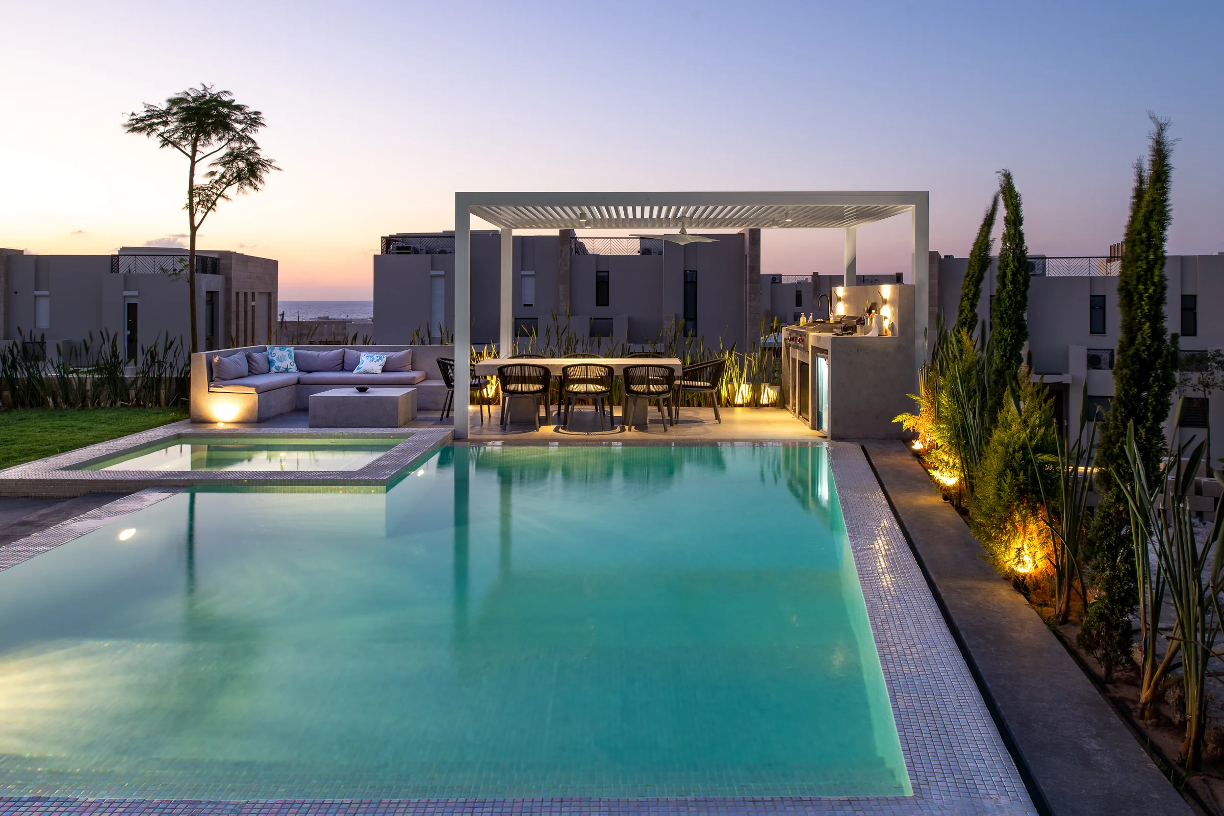 Dusk over the long pool, pergola lit from within.