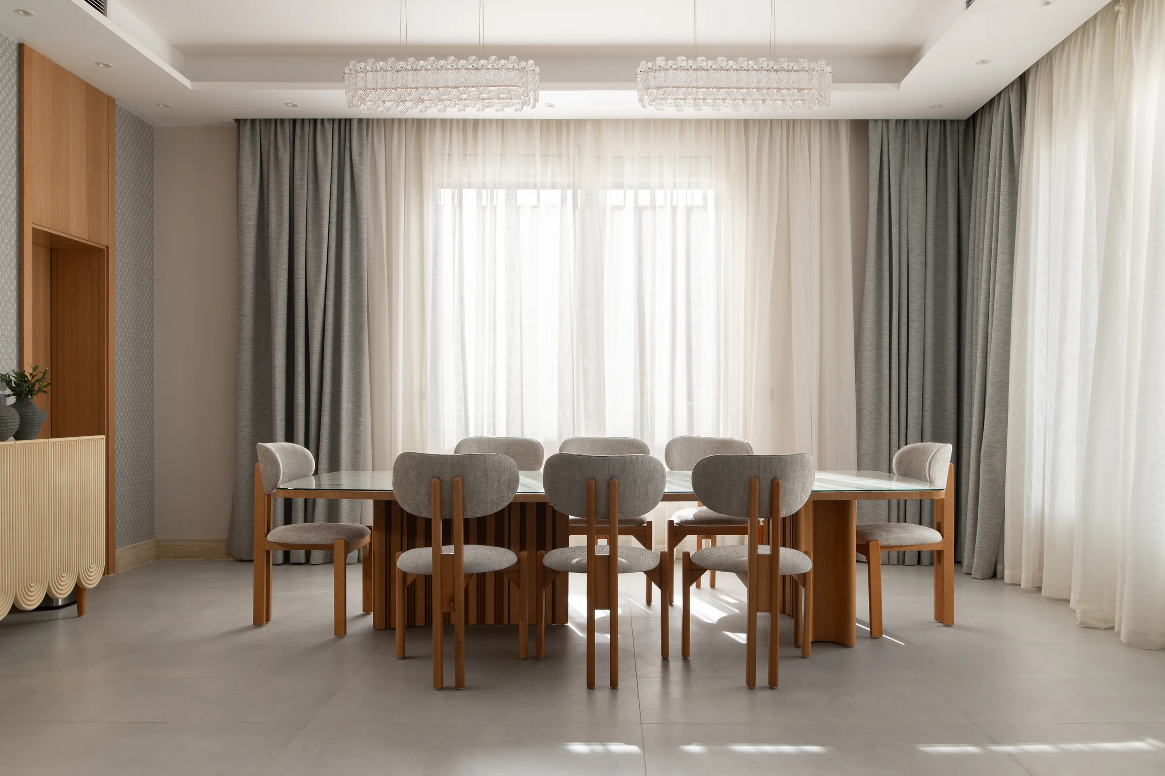 Dining table for eight under two crystal pendants, backlit by sheer curtains.